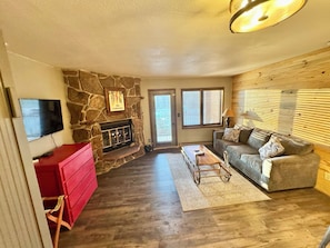 A living room with a stone fireplace, a television mounted on the wall, a red dresser, a grey sofa, and a wooden floor.