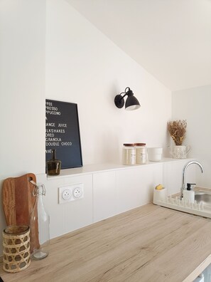 A modern kitchen with white cabinetry, a wooden countertop, a black wall-mounted lamp, and a white shelf with various items. - Studio Cosy - Plein Cœur Millau (Millau)
