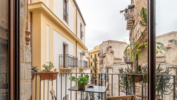 A narrow street with buildings on either side, balconies with potted plants, and a small table with a plant on it. - Loft Della Rosa by Wonderful Italy (Palermo)