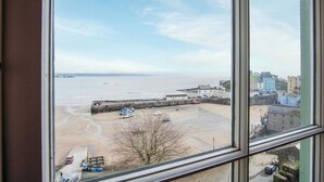 Vista costera a través de una ventana, que muestra una playa de arena, un muelle y edificios al fondo.