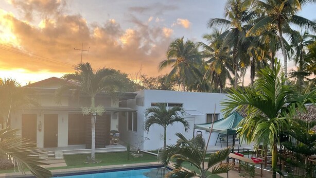 A swimming pool with a grassy area and palm trees, a white building with a garage, and a sunset in the background.