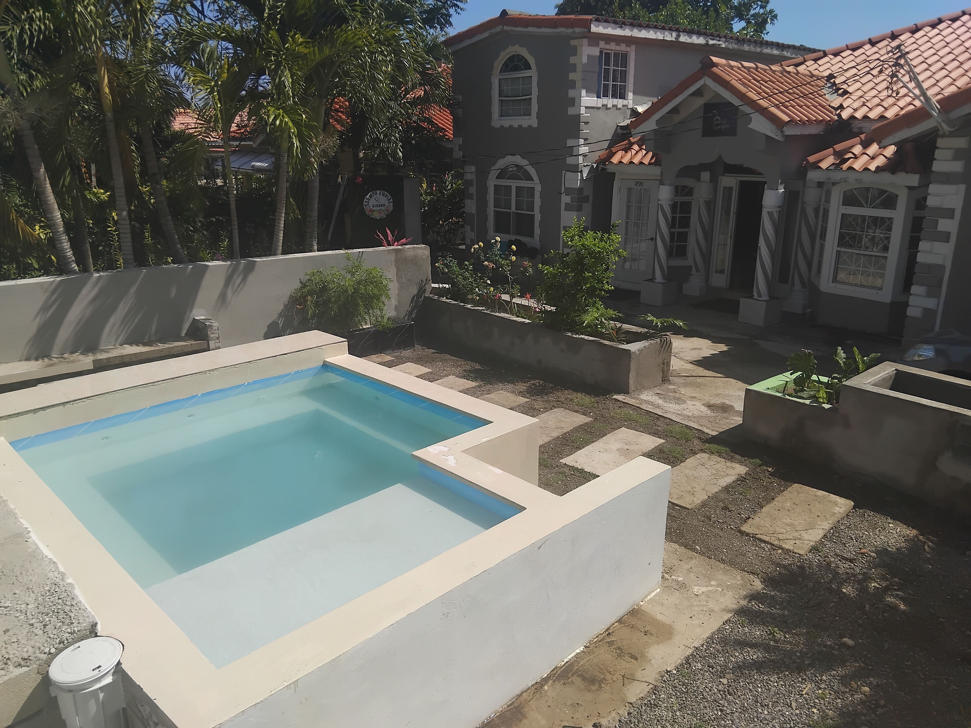 Une piscine avec une terrasse carrelée, entourée d’un jardin où poussent différentes espèces de plantes, et un bâtiment avec un toit en tuiles en arrière-plan.