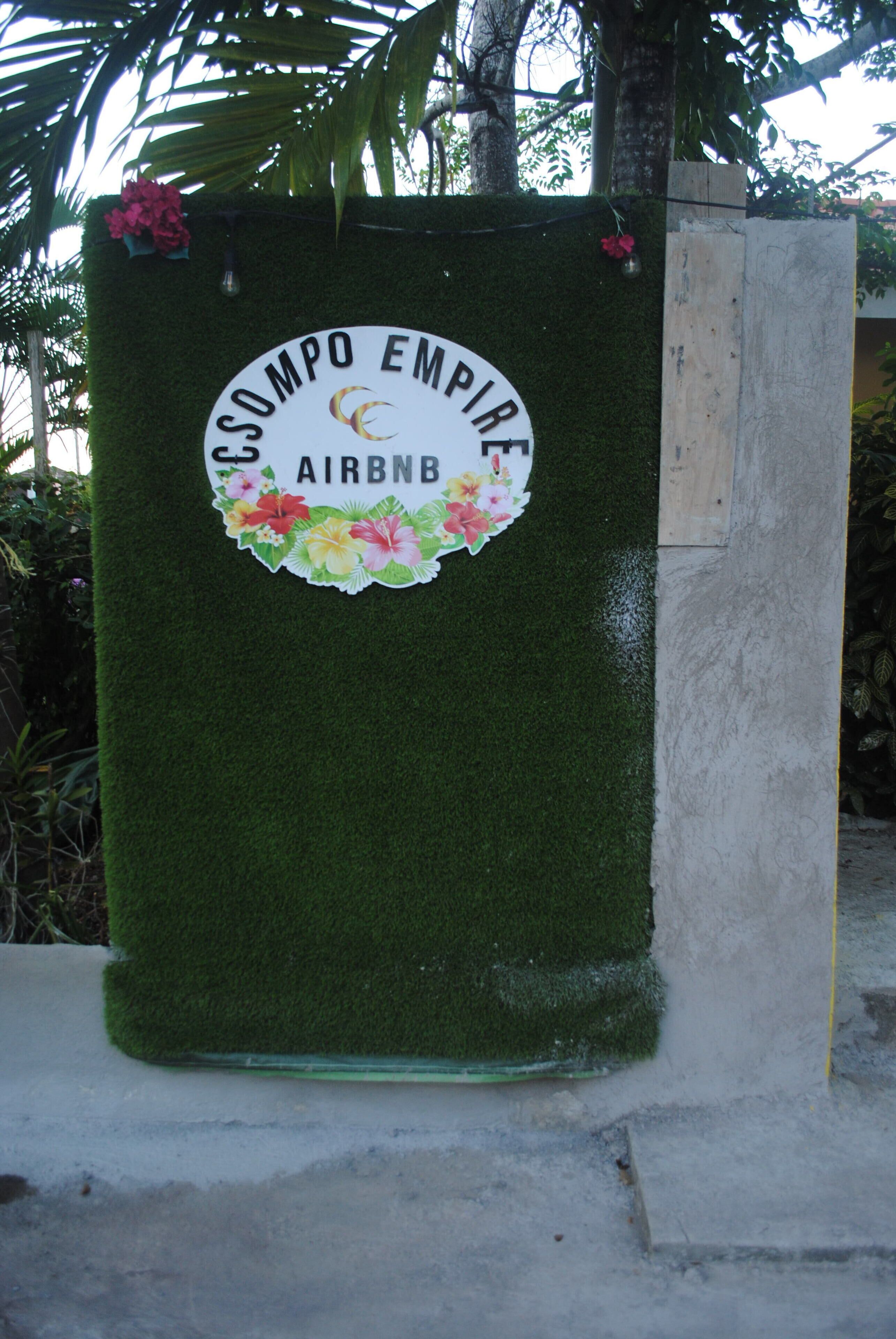 A green, textured wall with a circular sign featuring the Airbnb logo and the text "COMPO EMPIRE."