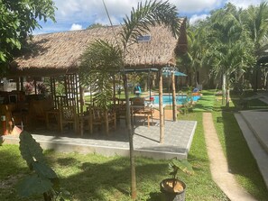A thatched-roof outdoor seating area with tables and chairs, a pool, and a well-maintained lawn. - Studio B, Loft (M&K’s Guest House) (Dauis)