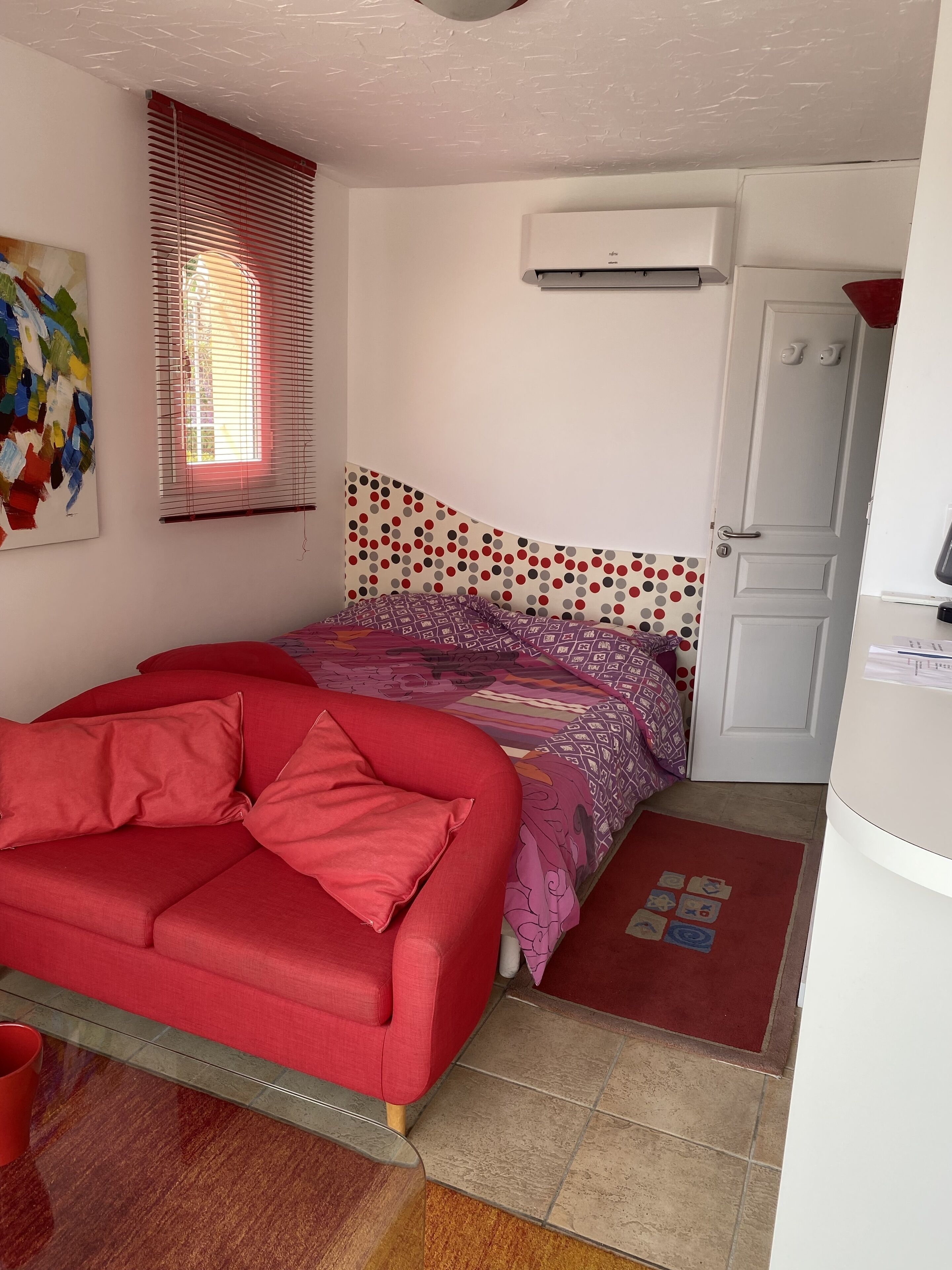 A bedroom with a red sofa, a patterned bedspread, a window with blinds, an air conditioning unit, and a door with a handle.