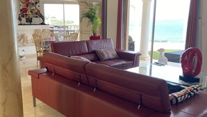 Een woonkamer met een grote rode bank, een eettafel met stoelen, een potplant en uitzicht op zee door grote ramen.