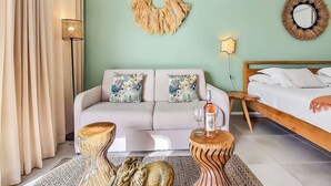 A modern hotel room with a light green wall, a sofa with tropical pillows, a wooden side table with a bottle and glass, a decorative elephant sculpture, and a bed with white linens.