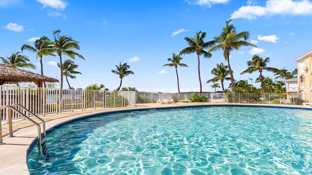 A clear blue pool with a white fence, palm trees, and a thatched-roof structure.