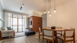 Een moderne woonkamer met een bank, een poef, en een eettafel met stoelen. Er is een televisie, een houten kast, en een groot raam met gordijnen.