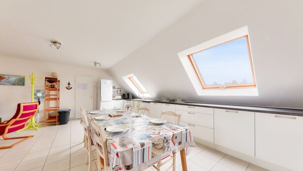 Un comedor amplio con una mesa preparada para cuatro personas, una cocina con armarios blancos y un tragaluz que proporciona luz natural.