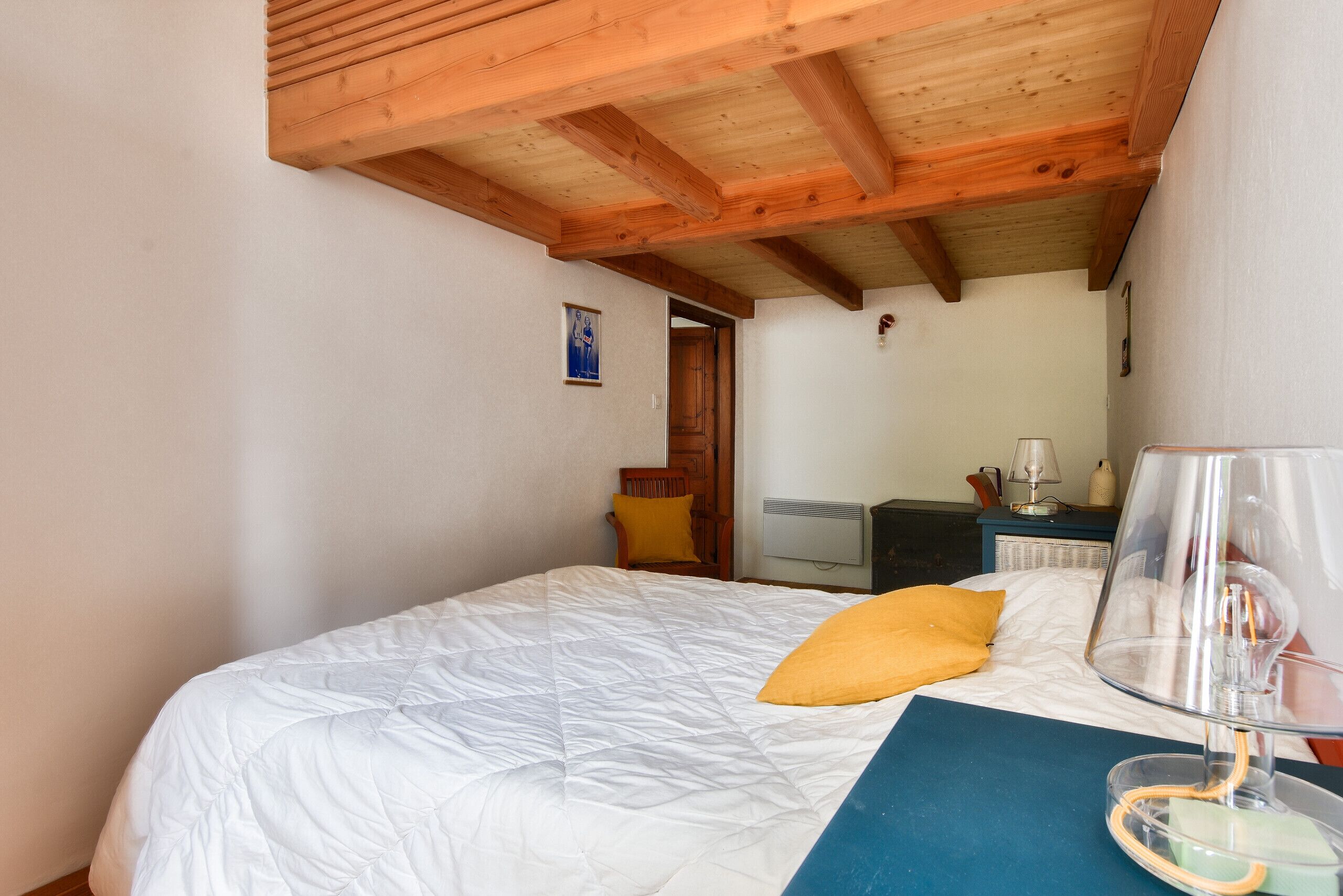 Une chambre avec un plafond en bois, un lit avec une literie blanche, un oreiller jaune, une table de chevet avec une lampe et un petit bureau également doté d’une lampe.