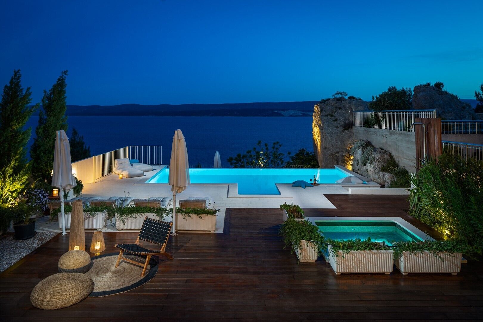 A luxurious outdoor pool area with a view of the sea, wooden deck, lounge chairs, and potted plants.