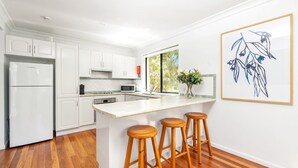 A modern kitchen with white cabinets, a built-in microwave, a coffee maker, and a large window with a view of greenery. There are three wooden stools at the kitchen island. - Sandpiper, Hyams Beach, Jervis Bay (Hyams Beach)
