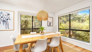 A dining area with a wooden table, white chairs, a hanging light fixture, and large windows with a view of trees. - Sandpiper, Hyams Beach, Jervis Bay (Hyams Beach)