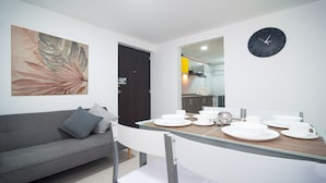 Un comedor moderno con una mesa de vidrio, sillas blancas y un sofá gris con cojines. Un reloj grande está colgado en la pared, y se ve una cocina al fondo.