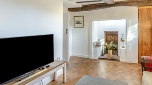 A living room with a large flat-screen TV on a wooden stand, a herringbone-patterned wooden floor, exposed wooden beams on the ceiling, and a doorway leading to a room with a brick fireplace. - 2 bedroom accommodation in Much Hadham (Much Hadham)