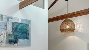 Een kamer met zichtbare houten balken, een hangende geweven lamp en een groot abstract schilderij aan de muur.