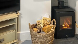 Een woonkamer met een televisie op een houten standaard, een zwarte houtkachel en een rieten mand vol met houtblokken.