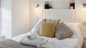 Een net opgemaakt bed met kussens en een opgevouwen handdoek, een nachtkastje met boeken en een kleine plant, en wandlampen.