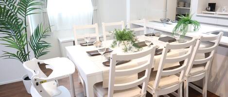 Un área de comedor moderna con una mesa y sillas blancas, una planta en maceta y una cocina al fondo.