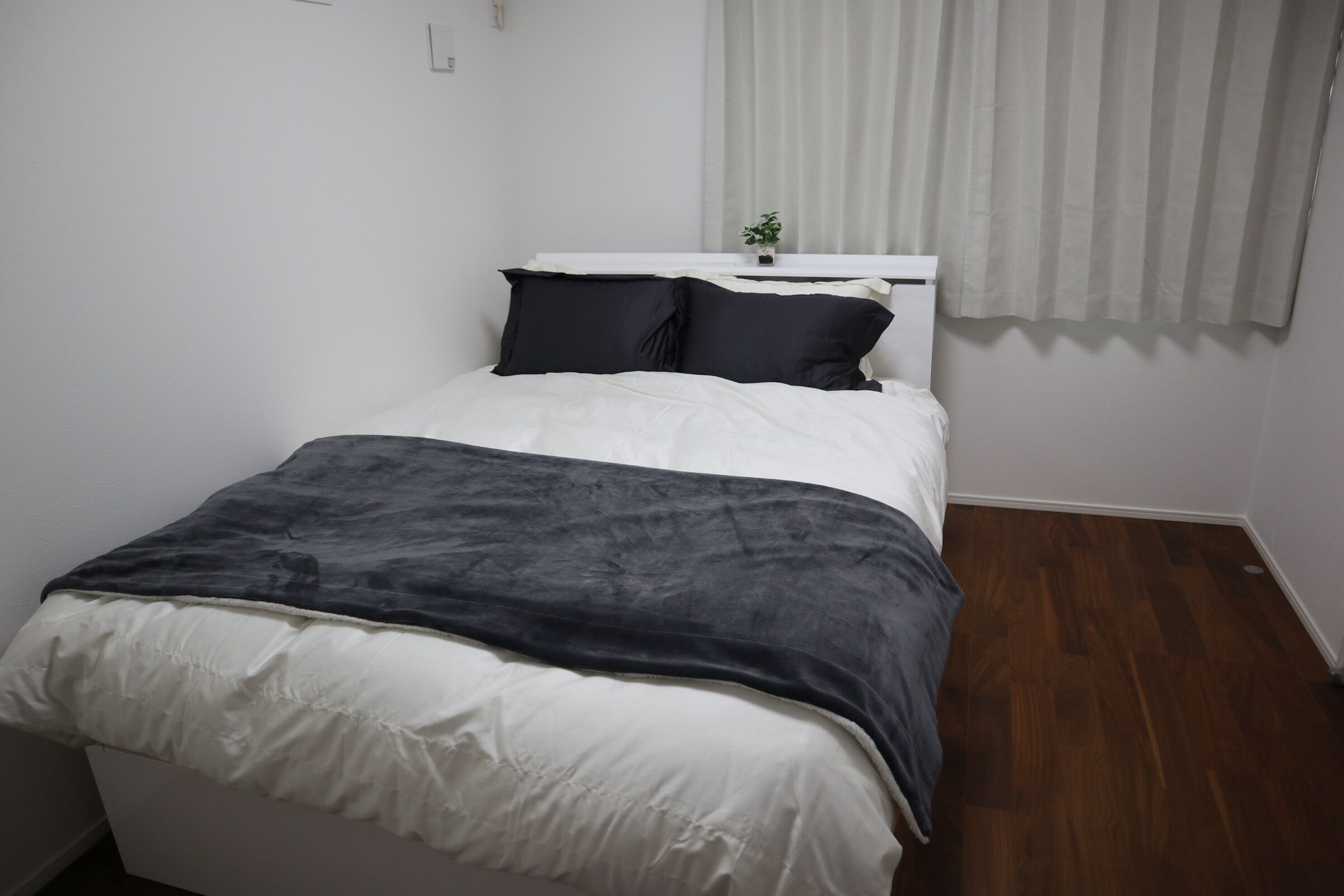 A bedroom with a bed, white walls, a window with blinds, and a small plant on the windowsill.