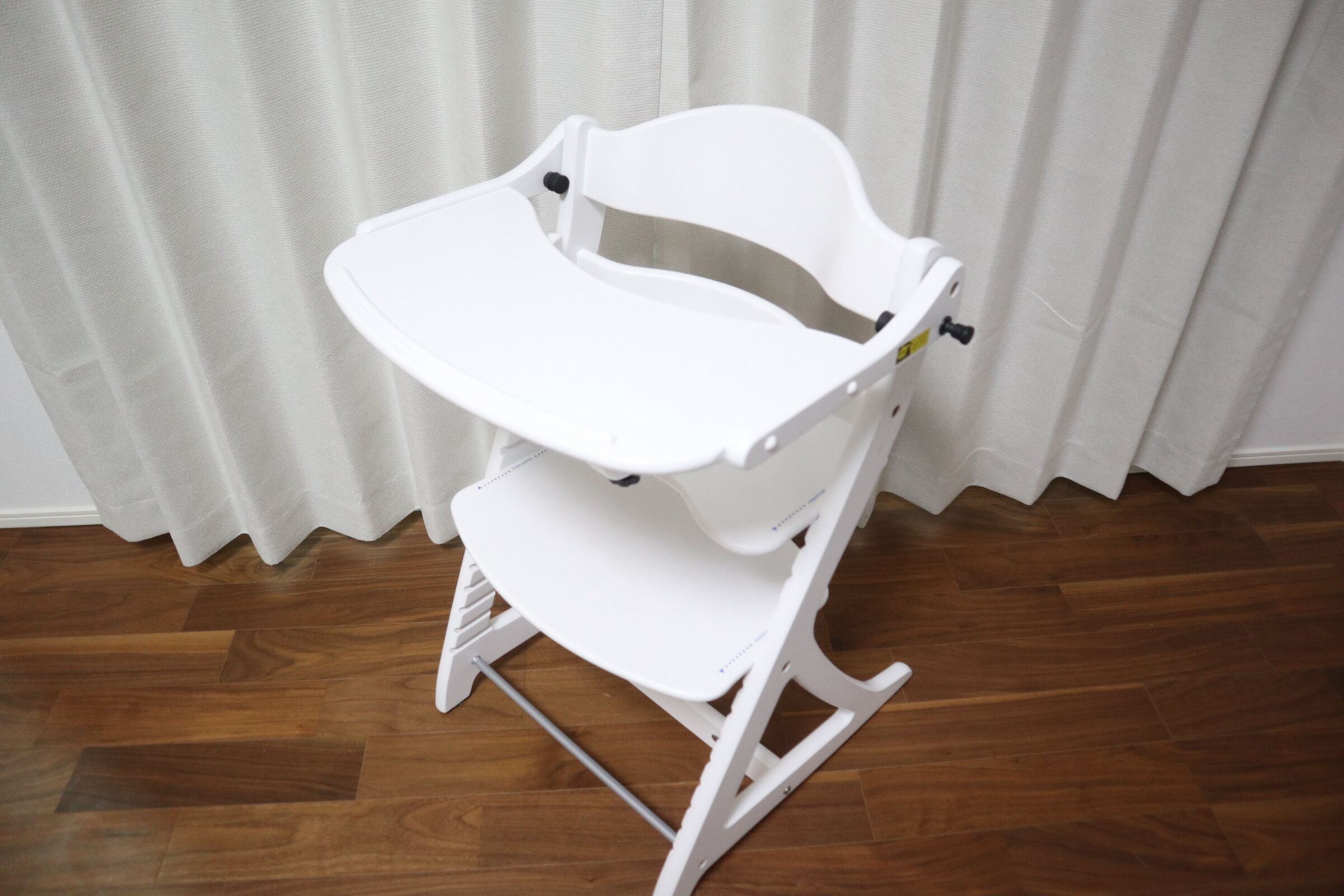 A white baby rocker with a tray, placed on a wooden floor against a white curtain backdrop.