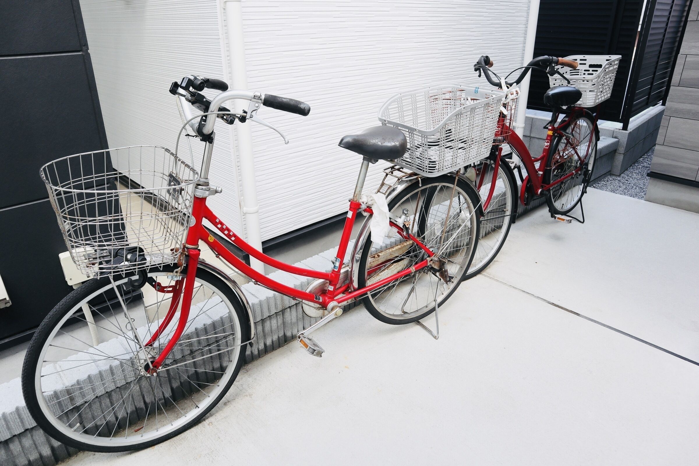 A red bicycle with a basket and a white basket attached to the back, parked on a concrete surface.