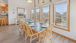 Ruokailutilassa on suuri pöytä, johon on katettu kuudelle henkilölle. Pöydän ympärillä on puisia tuoleja, ja suurista ikkunoista avautuu näkymä asuinalueelle.