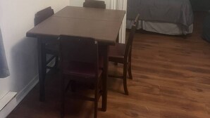 Une petite pièce avec une table et des chaises en bois, un lit recouvert d’une couverture grise et un téléviseur posé sur un meuble.