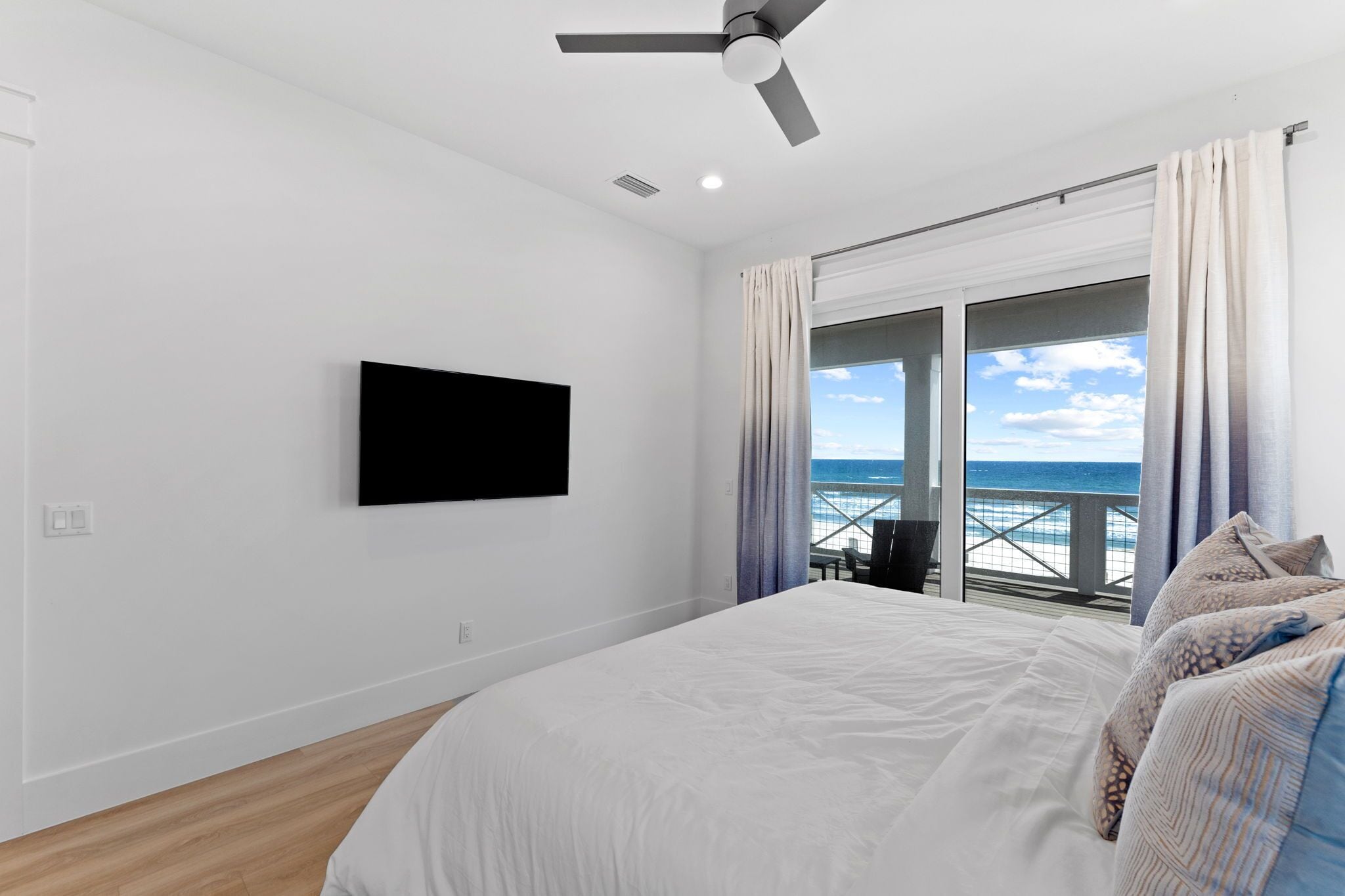Et soveværelse med seng, ventilator, fjernsyn monteret på væggen og en balkon med havudsigt.