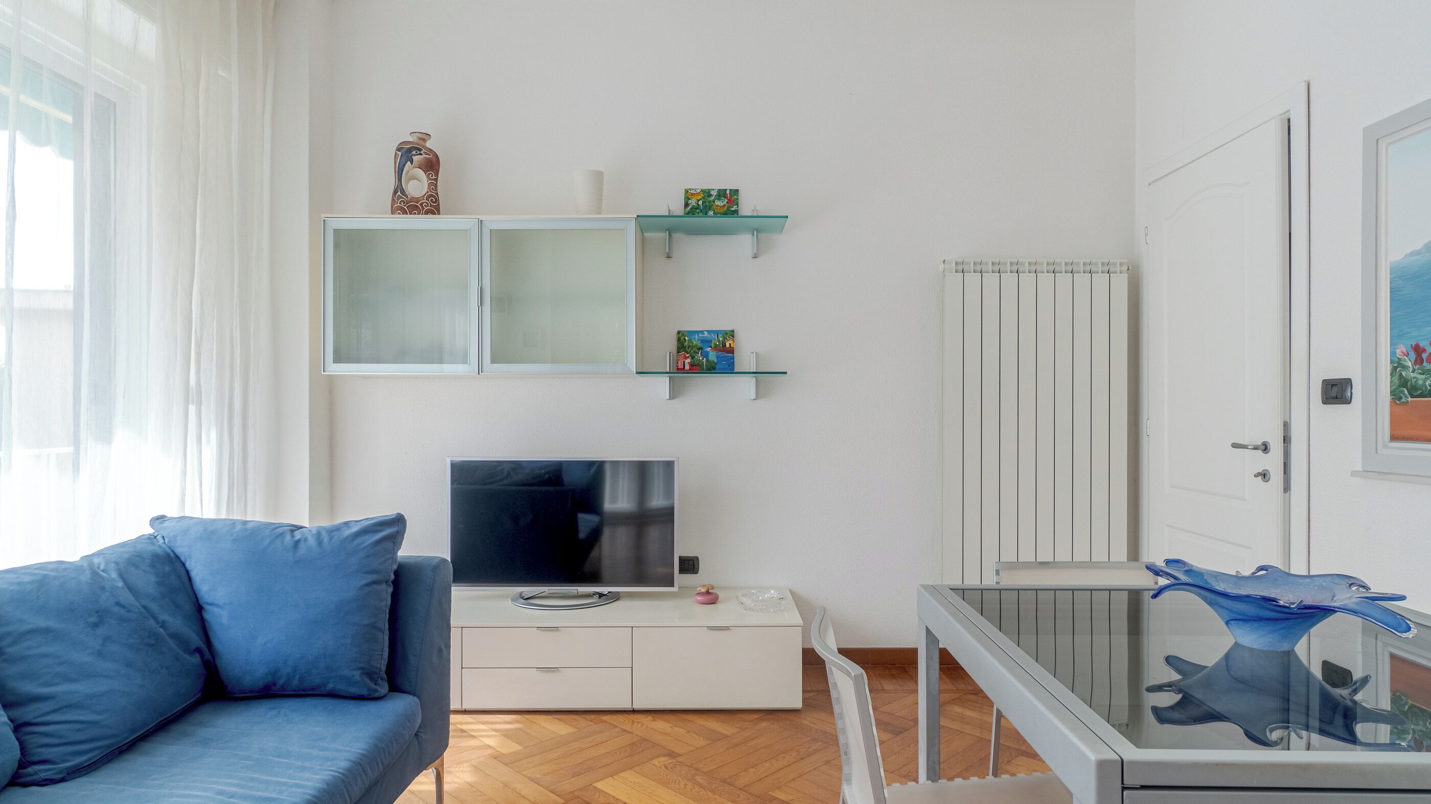 A room with a blue sofa, a TV on a white cabinet, a glass table with a blue sculpture, and a door with a painting.