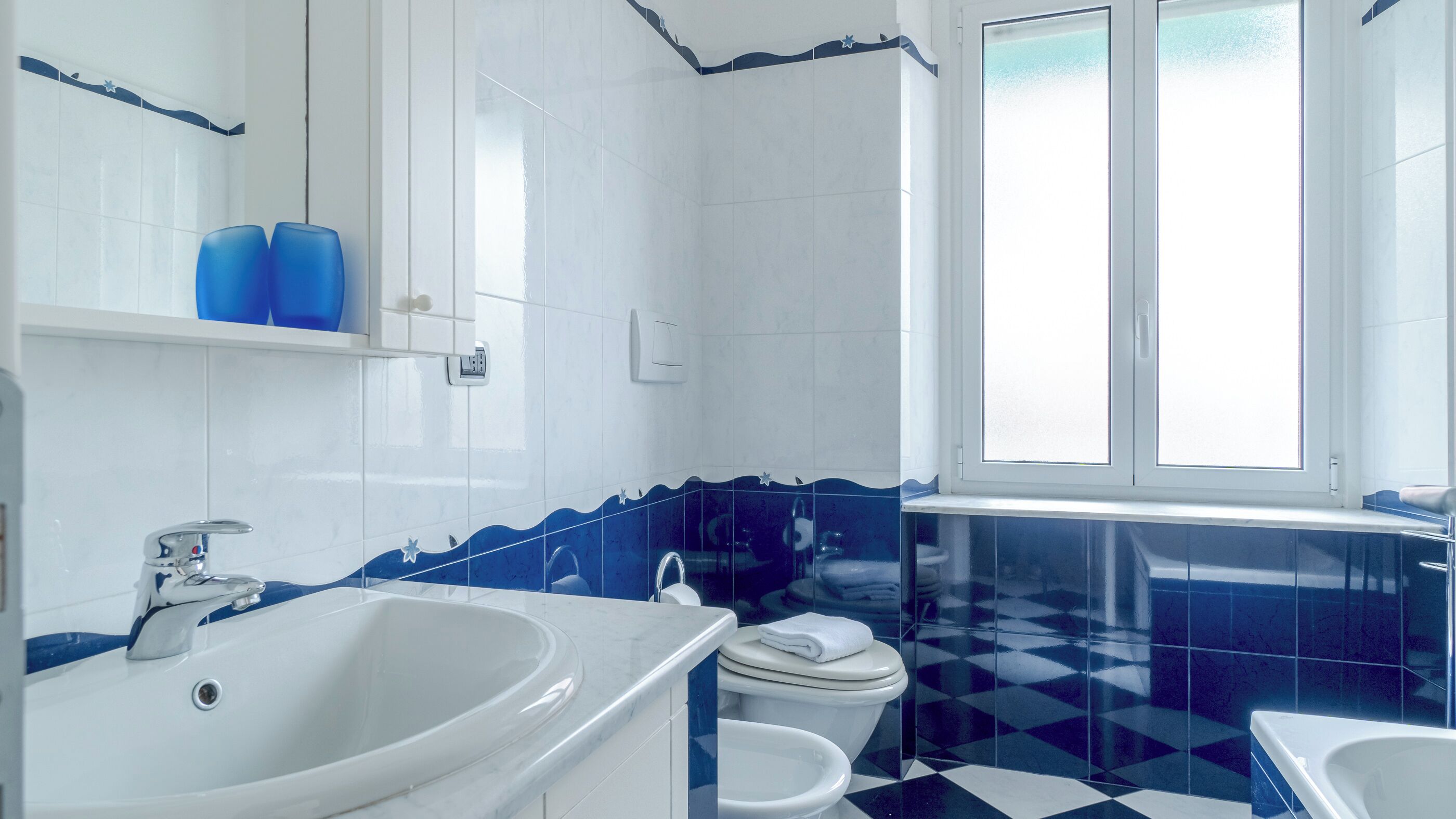 A bathroom with a white sink, a toilet, and a bathtub. There is a window with blinds, and the walls are tiled with a blue and white pattern.