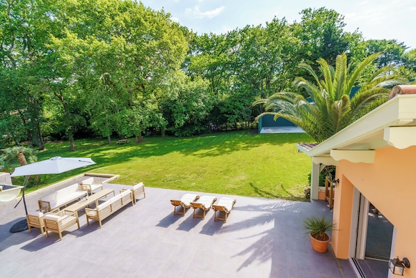 Un espace patio aménagé avec du mobilier d’extérieur, un grand parasol et une vue sur un jardin luxuriant, avec des arbres et un palmier.