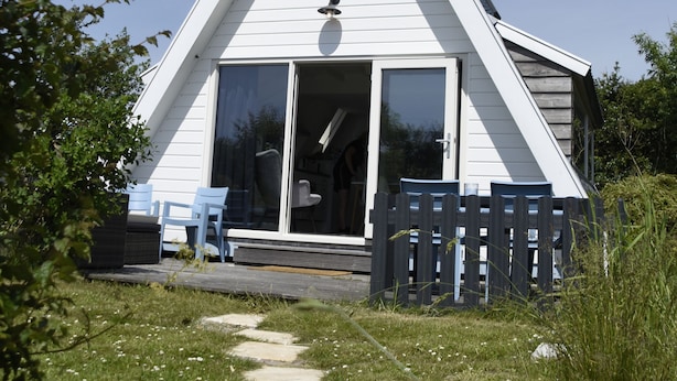 Un petit bâtiment blanc, doté d’un toit triangulaire, avec une porte d’entrée coulissante en verre, un petit porche avec une clôture en bois et un chemin menant à la porte.