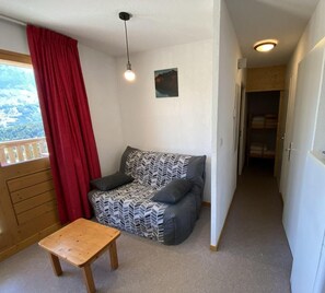 Ein kleiner, gemütlicher Raum mit einer Couch, einem Holztisch und Blick durch ein Fenster mit roten Vorhängen.