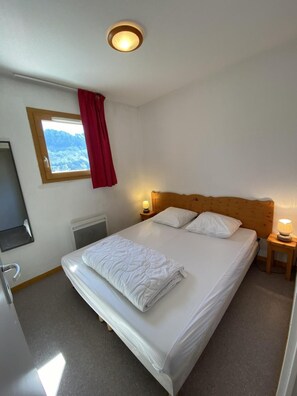 A single bed with a wooden headboard, a bedside table with a lamp, a window with a view of mountains, and a red curtain.