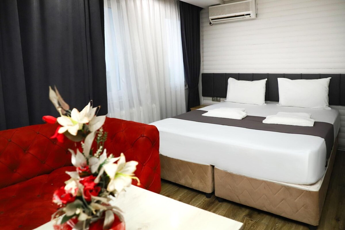 Una habitación de hotel con una cama, un sofá rojo, una mesa blanca y un florero con flores.