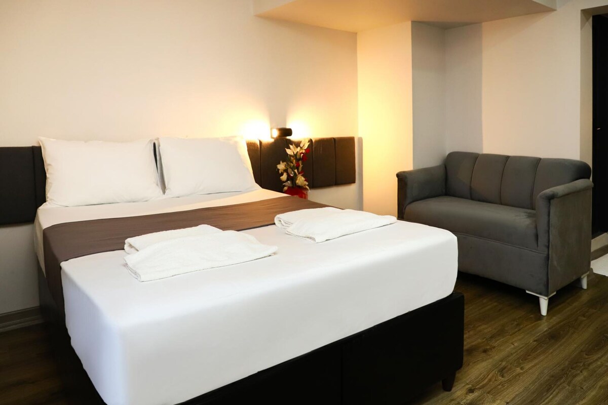 Una habitación de hotel con una cama, una mesita de noche con un florero y un sillón gris.