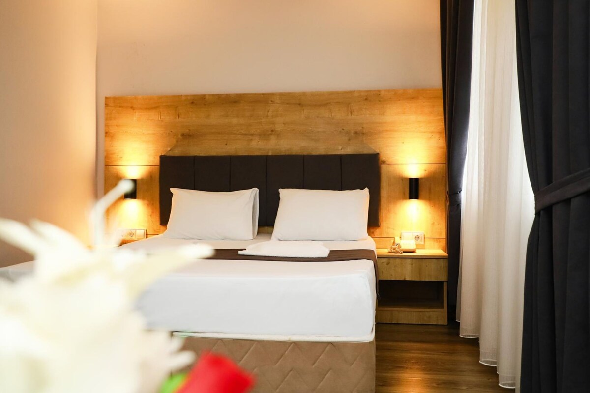 Habitación de hotel con cabecera de madera, dos mesitas de noche con lámparas, cama con ropa blanca y vista a una cortina.