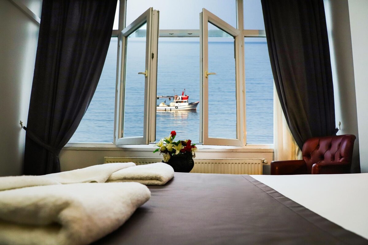 Habitación de hotel con cama, ventana con cortinas, un jarrón con flores y vista al mar con un barco.