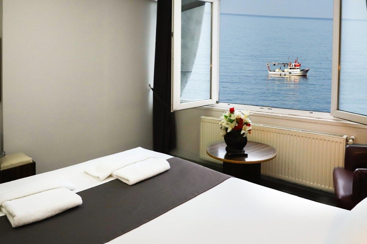 Habitación de hotel con una cama, ventana con vista al mar, una mesita con un florero y una silla.