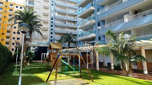 A residential building with multiple balconies, a playground with swings and slides, and palm trees.