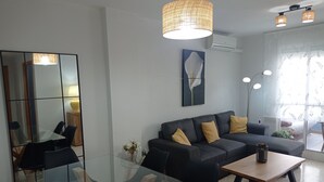 A modern living room with a glass table, dark sofa with yellow cushions, a large mirror, and a hanging light fixture.