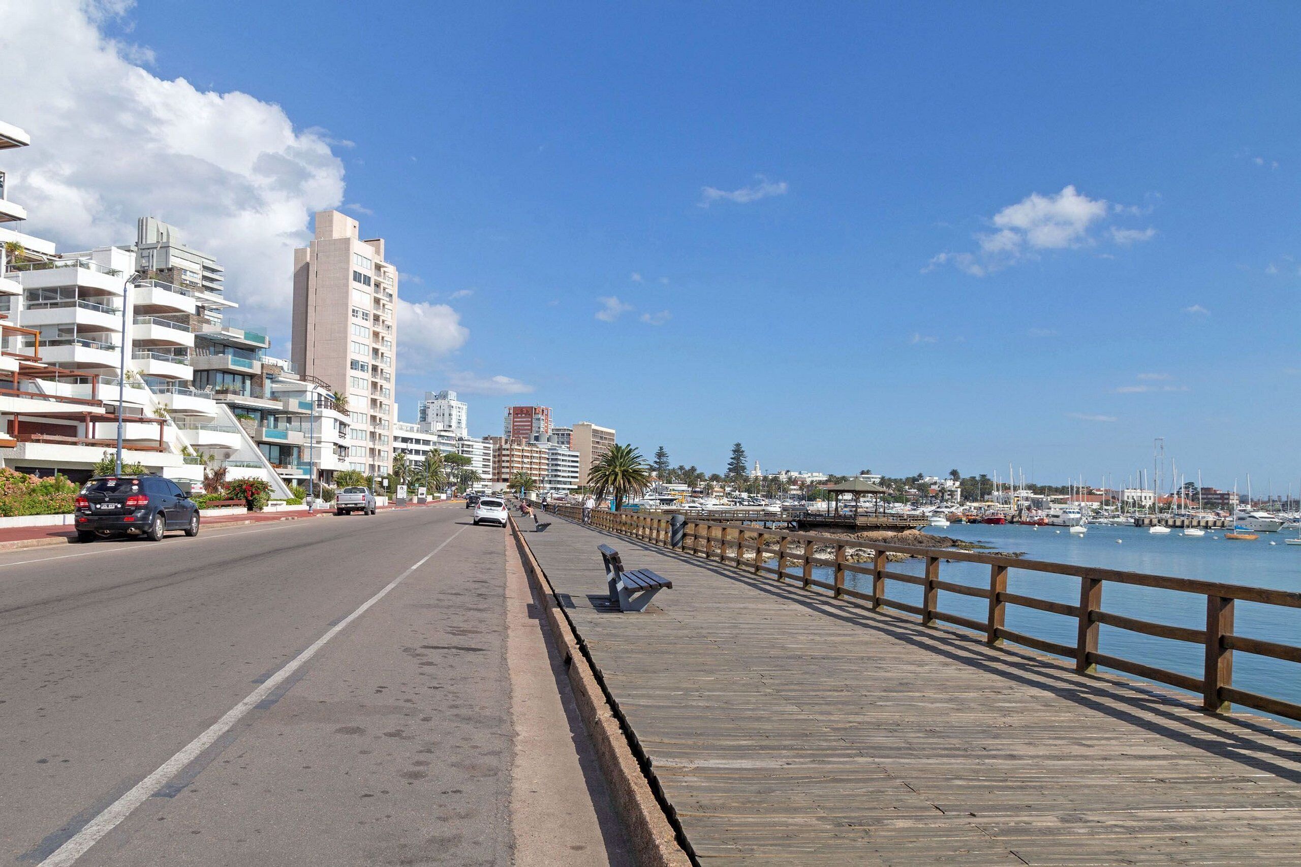 Una carretera costera con un paseo de madera, vehículos y edificios al fondo.