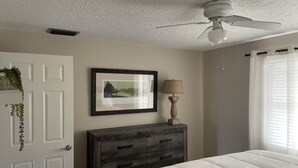 Une chambre à coucher avec un lit, une commode, un ventilateur de plafond, un tableau encadré et une fenêtre avec des rideaux.