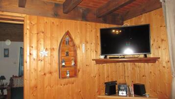 Une pièce aux murs lambrissés, avec un téléviseur fixé sur une poutre en bois, une petite étagère ornée de figurines et un haut-parleur posé sur une table en bois.