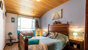 Ein Schlafzimmer mit Holzbalkendecke, einem Bett mit floralem Bettbezug, einem Nachttisch mit Lampe und einem Fenster mit Blick nach draußen.