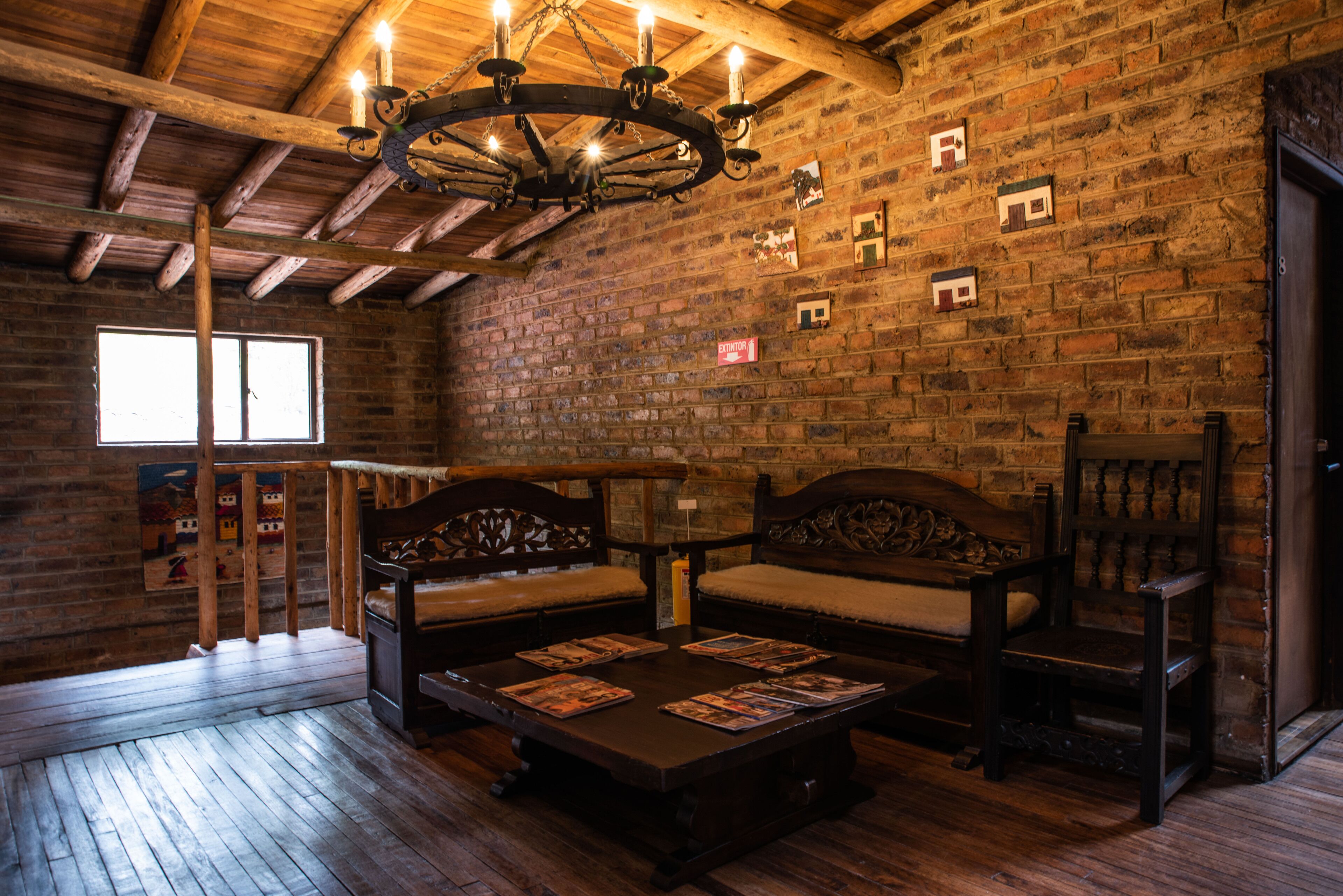 Una habitación rústica con vigas de madera a la vista, pared de ladrillo y suelo de madera. Cuenta con un banco de madera con cojines, una mesa de madera con revistas y una silla de madera.