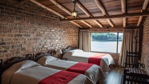 Una habitación con dos camas, techo de madera, pared de ladrillo, ventana con vista a un lago y un banco.