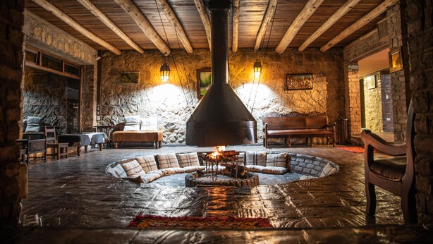 Un interior rústico con paredes de piedra, vigas de madera, un hogar central y asientos alrededor.